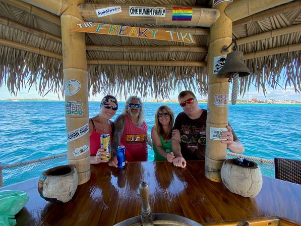 Four smiling adults at a bamboo tiki bar on a boat, holding canned drinks under a thatched roof with bright turquoise ocean and distant shoreline — sunny tropical vacation vibe