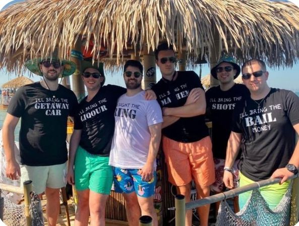 Six friends in sunglasses and swim trunks at a beachside tiki hut, wearing novelty matching t-shirts with playful slogans, colorful shorts and straw hats, ocean visible behind them.