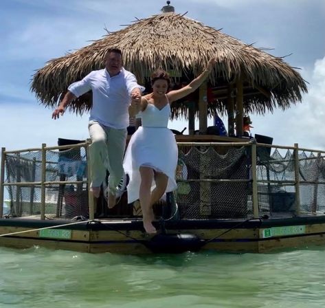 Joyful bride and groom holding hands as they leap from a thatched-roof tiki boat into shallow turquoise tropical water under a sunny blue sky.