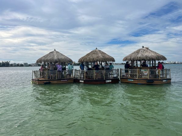 Three thatched-roof floating tiki-bar pontoons clustered on green bay water, people gathered under the huts, cloudy blue sky and a distant shoreline of low buildings.