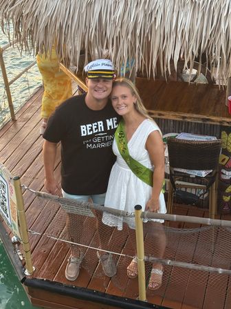 Smiling couple on a tropical wooden dock under a thatched tiki roof — man in a captain's hat and 'Beer Me I'm Getting Married' shirt, woman in a white dress wearing a green bride-to-be sash, water and tiki bar details visible.