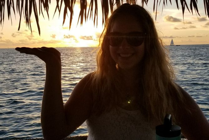 Silhouette of a person in sunglasses under thatched palm fronds, playfully holding the setting sun over a tropical ocean at sunset with a sailboat on the horizon.