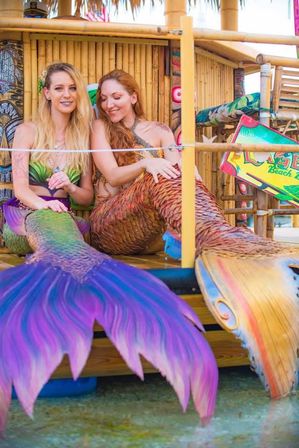 Two mermaid performers with colorful iridescent tails (purple-green and copper-gold) relaxing in a bamboo tiki hut at a tropical beachside setting