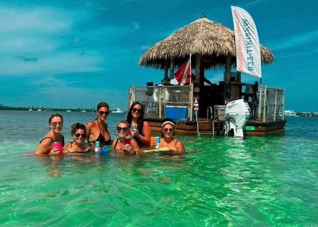 Six friends in swimsuits holding colorful drinks in shallow, clear turquoise water beside a thatched-roof floating tiki hut with an outboard motor under a bright blue sky in a tropical bay