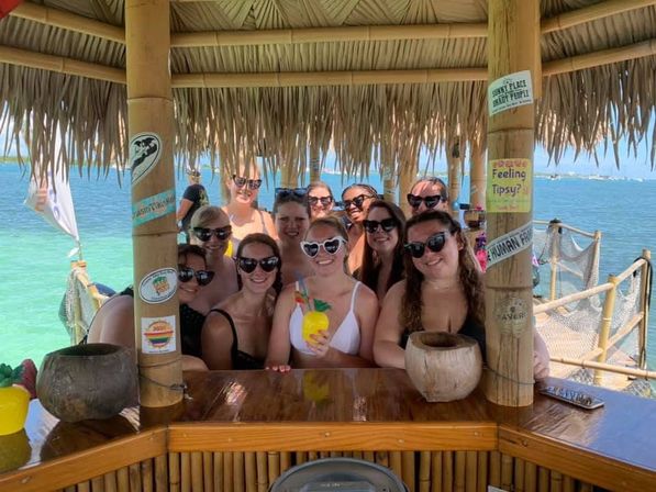 Smiling group of women in sunglasses at a bamboo tiki bar on a sunny turquoise ocean dock, holding tropical cocktails under a thatched roof
