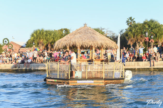 Tiki-style floating bar boat cruising on blue water past a packed waterfront seawall with palm trees and spectators on a sunny day