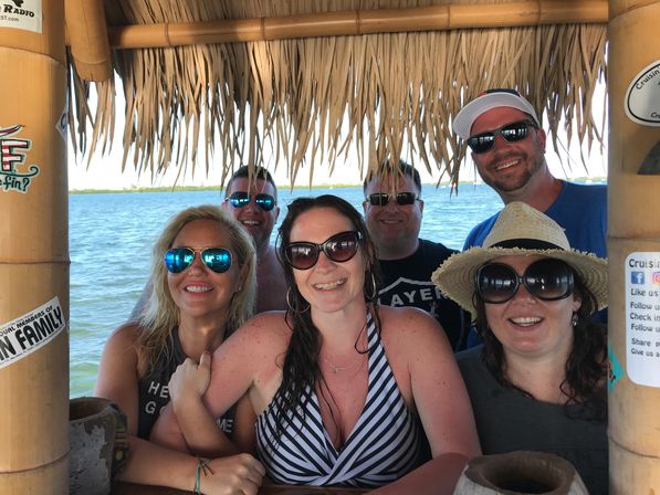 Six friends smiling under a bamboo thatched tiki hut at a sunny waterfront bar, wearing sunglasses, swimsuits and a straw hat with blue bay water and distant shoreline behind them.