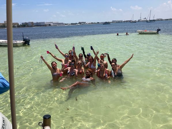Group of people in swimsuits posing and cheering in shallow clear turquoise bay, holding drinks with small boats and a coastal skyline in the background on a sunny day
