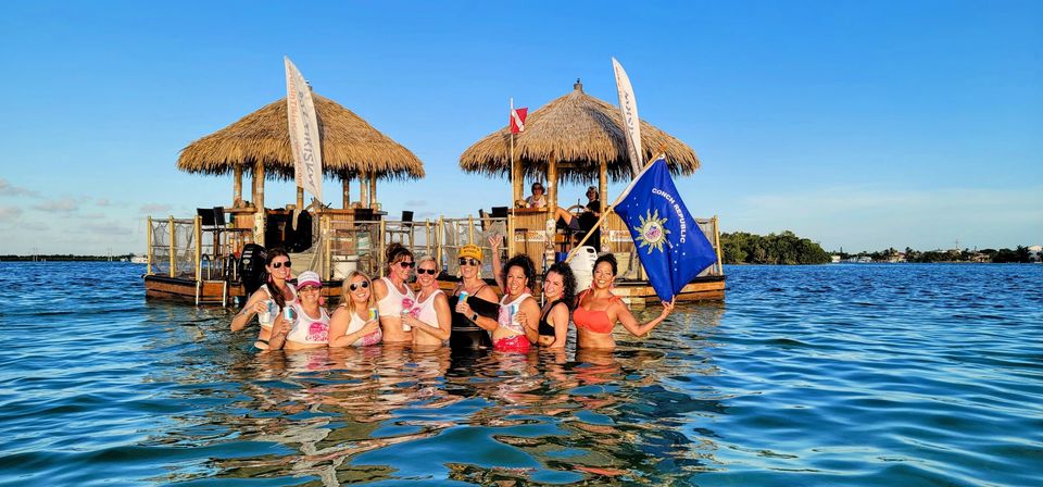 Group of friends in swimsuits wading in clear blue coastal water in front of a floating tiki-bar raft with thatched roofs and flags under a sunny sky
