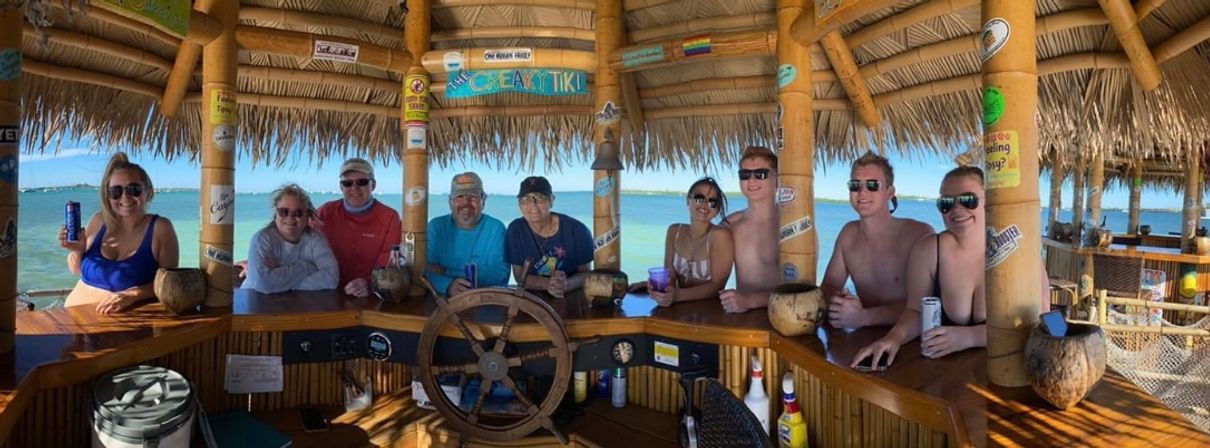 Smiling group at a thatched-roof waterfront tiki bar with a wooden ship wheel, holding drinks over turquoise ocean on a sunny tropical day