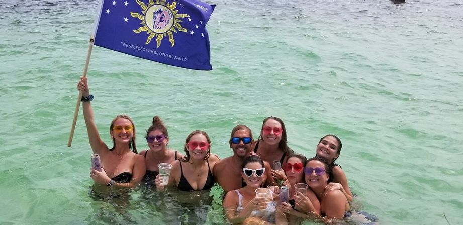 Group of eight people waist-deep in clear turquoise ocean, smiling and holding drinks at a beach party while one person waves a blue sun-themed flag and several wear colorful sunglasses.