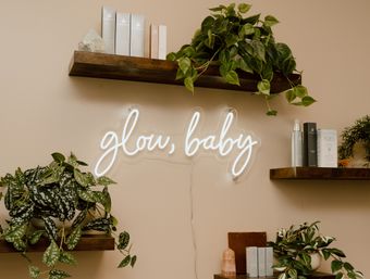 White neon sign reading "glow, baby" on a beige wall above wooden floating shelves with trailing indoor plants, skincare products, and a rose quartz crystal — cozy beauty and wellness display.