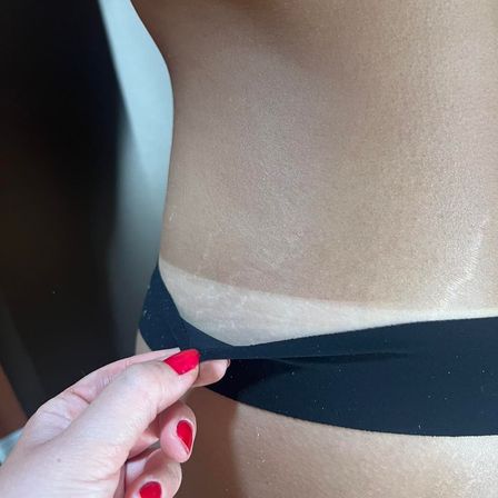 Close-up of sun-kissed hip with a distinct bikini tan line, hand with red nail polish pulling down a black swimsuit strap — summer beach vacation vibe