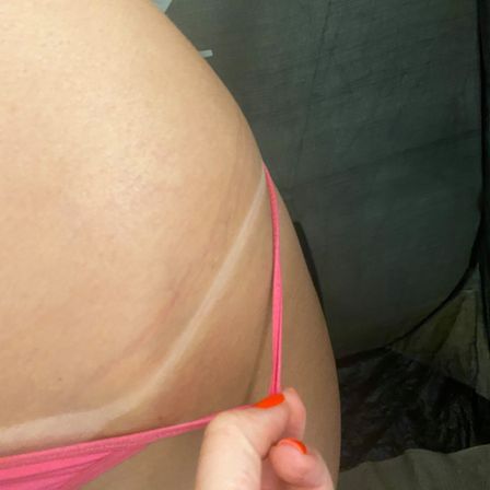 Close-up of hip showing a white tan line as a hand with orange-painted nails pulls a pink bikini strap, summer swimwear detail