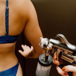 Close-up of a person in a blue patterned bikini receiving a spray tan on their back, with a technician’s painted nails holding a spray-tanning gun.