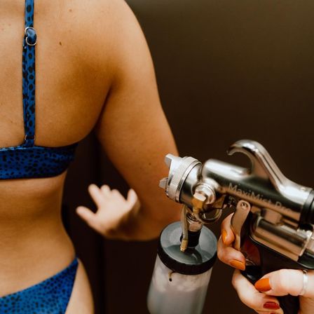 Close-up of a person in a blue patterned bikini receiving a spray tan on their back, with a technician’s painted nails holding a spray-tanning gun.
