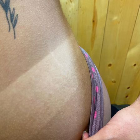 Close-up of a hip with a distinct bikini tan line, pink-dotted underwear edge, small floral tattoo and wood-panel background