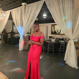 Smiling woman in a red off-shoulder evening gown posing on a dance floor inside a decorated banquet hall with sheer white drapes, crystal chandelier and tables set for a formal event.