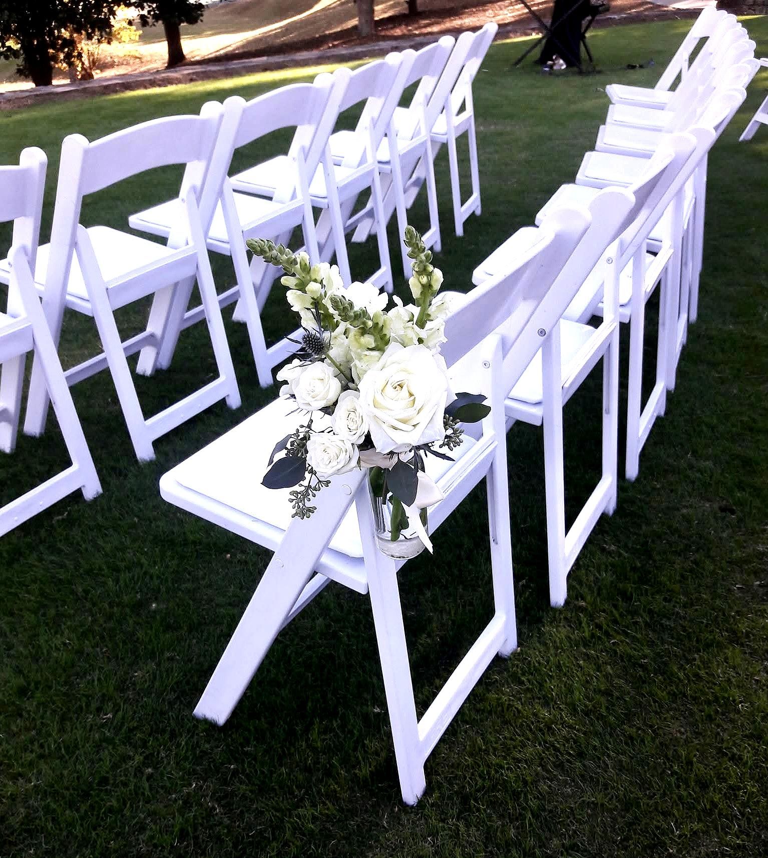 Outdoor wedding aisle with rows of white folding chairs on green lawn, close-up of a white rose and greenery bouquet tied to the end chair