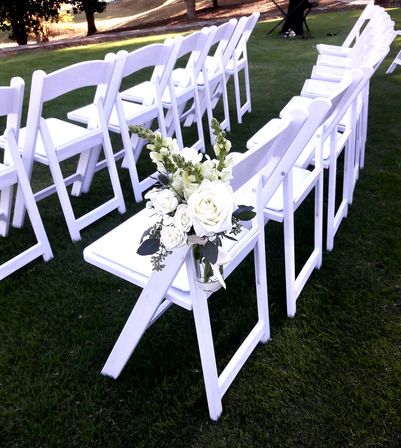Outdoor wedding aisle with rows of white folding chairs on green lawn, close-up of a white rose and greenery bouquet tied to the end chair
