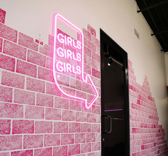 Playful pink neon sign reading "GIRLS GIRLS GIRLS" with an arrow mounted on a pink brick-patterned interior wall beside a black glass door entrance.