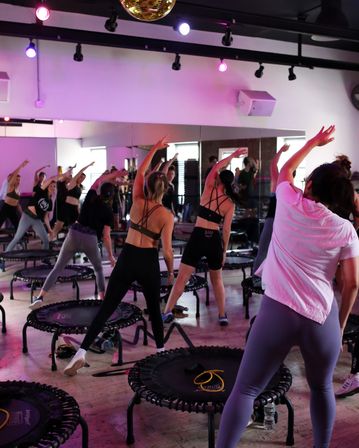 High-energy group trampoline rebounder workout in a mirrored indoor fitness studio with purple lighting and a disco ball, participants stretching and bouncing during a group fitness class.