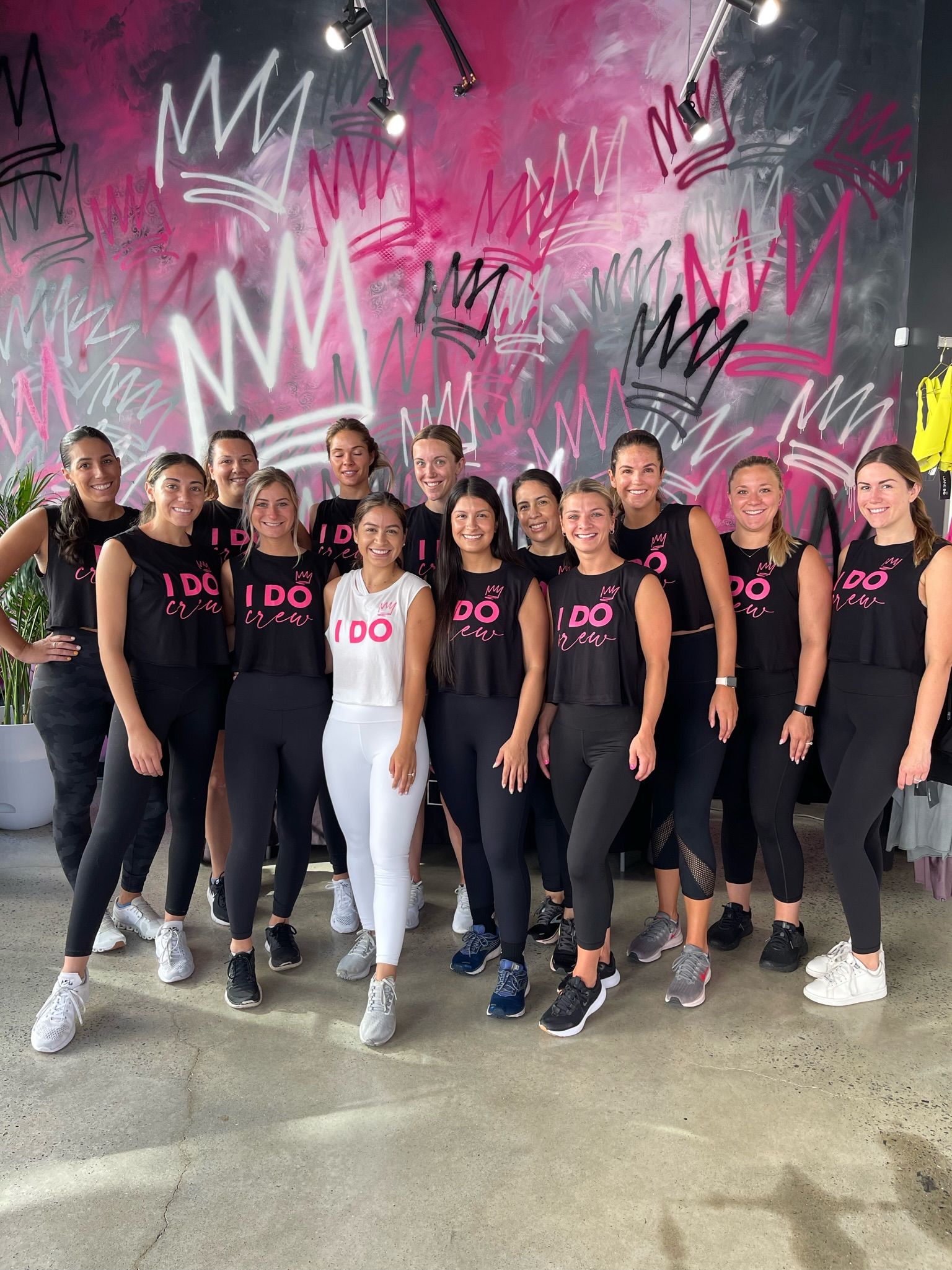 Group of women in matching "I DO" workout tops and leggings posing in a boutique fitness studio with a vibrant pink and black crown graffiti mural — bachelorette/bridal workout group photo.