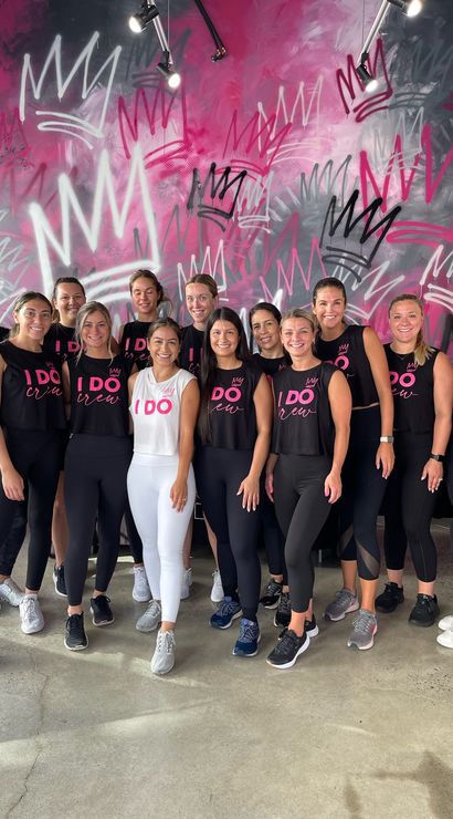 Group of women in matching "I DO" workout tops and leggings posing in a boutique fitness studio with a vibrant pink and black crown graffiti mural — bachelorette/bridal workout group photo.
