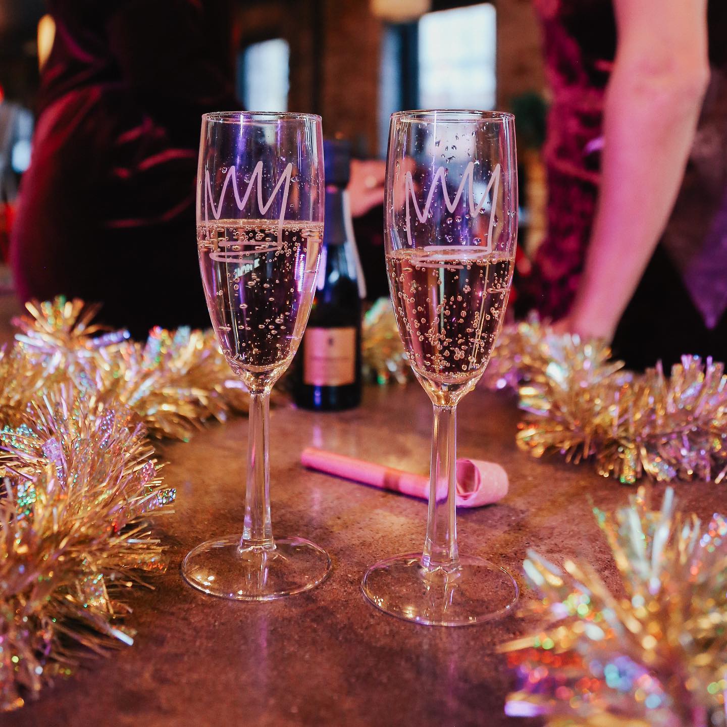 Two bubbly champagne flutes with crown designs on a festive indoor party table surrounded by gold tinsel, a bottle and a pink party blower — New Year’s celebration vibe.