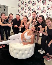 Group of women in athletic wear celebrating after a group workout in a boutique fitness studio — one lounging on a white tufted ottoman while classmates point and laugh in front of pop-art wallpaper and lockers.