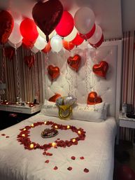 Romantic hotel room decorated for newlyweds with red and white balloons and red foil heart balloons above a white tufted headboard, bed topped with a heart of red rose petals, battery tealight candles and “Just Married” pillows.