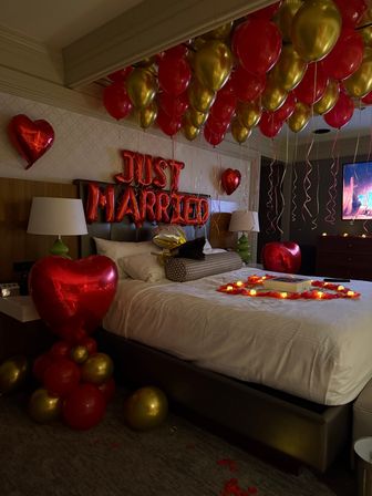 Romantic hotel room decorated for newlyweds with red "JUST MARRIED" foil letters, dozens of red and gold helium balloons on the ceiling, heart-shaped balloons, rose petals and LED candles on the bed.