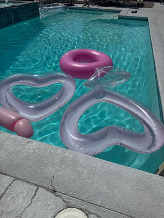 Sunlit outdoor swimming pool with clear turquoise water, a bright pink donut float and translucent heart-shaped inflatables drifting near a textured concrete pool deck—sparkling summer vibes.
