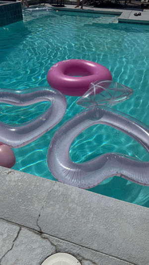 Sunlit outdoor swimming pool with clear turquoise water, a bright pink donut float and translucent heart-shaped inflatables drifting near a textured concrete pool deck—sparkling summer vibes.