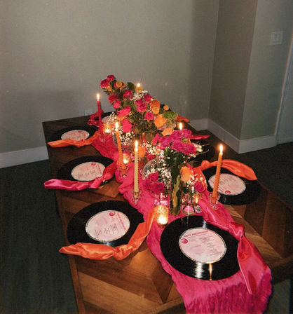 Indoor dining tablescape with pink and orange floral centerpiece, lit taper candles, disco balls and vinyl record placemats tied with satin ribbons — vibrant party decor.