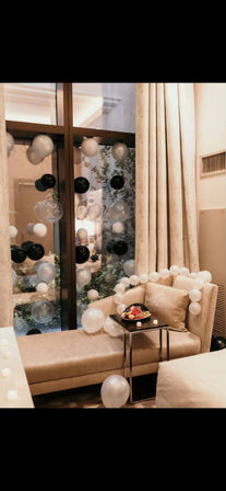 Cozy beige hotel suite window seat decorated for a celebration with black, white and clear balloons, a balloon garland draped over the chaise, and a small side table holding a fruit platter and candles.