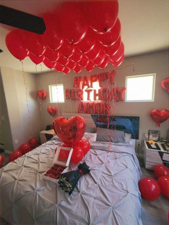 Bedroom staged for a romantic birthday surprise with dozens of red helium balloons on the ceiling, red foil "HAPPY BIRTHDAY" letters on the wall, heart-shaped balloons, and a bed with a bouquet and box of red roses.
