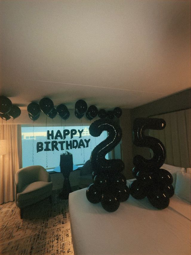 Room interior decorated for a 25th birthday with large black 2 and 5 balloons, 'HAPPY BIRTHDAY' window letters, clusters of black helium balloons and cozy seating by a sunlit window