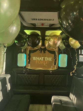 Party bus interior with black leather seating, gold and black balloons floating from the ceiling, teal window lights and a festive "What the Forty!" birthday banner beneath an emergency exit sign.