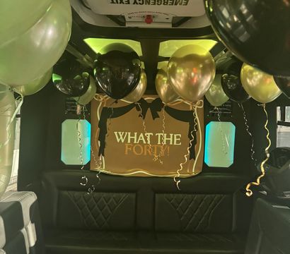 Party bus interior with black leather seating, gold and black balloons floating from the ceiling, teal window lights and a festive "What the Forty!" birthday banner beneath an emergency exit sign.