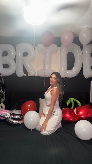 Bride-to-be kneeling on a bed in a decorated bedroom with large white 'BRIDE' letter balloons overhead and pink, red and white balloons, including heart-shaped foil balloons