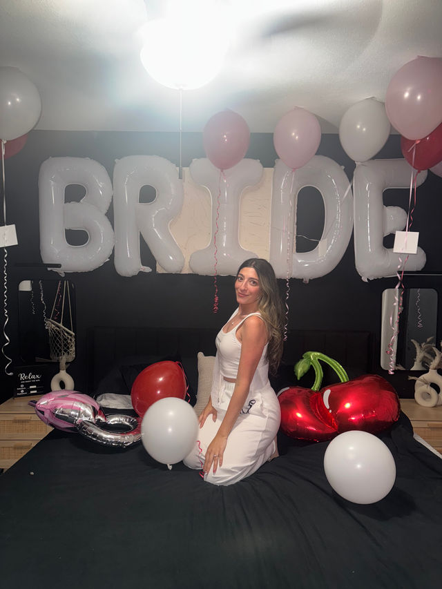 Bride-to-be kneeling on a bed in a decorated bedroom with large white 'BRIDE' letter balloons overhead and pink, red and white balloons, including heart-shaped foil balloons