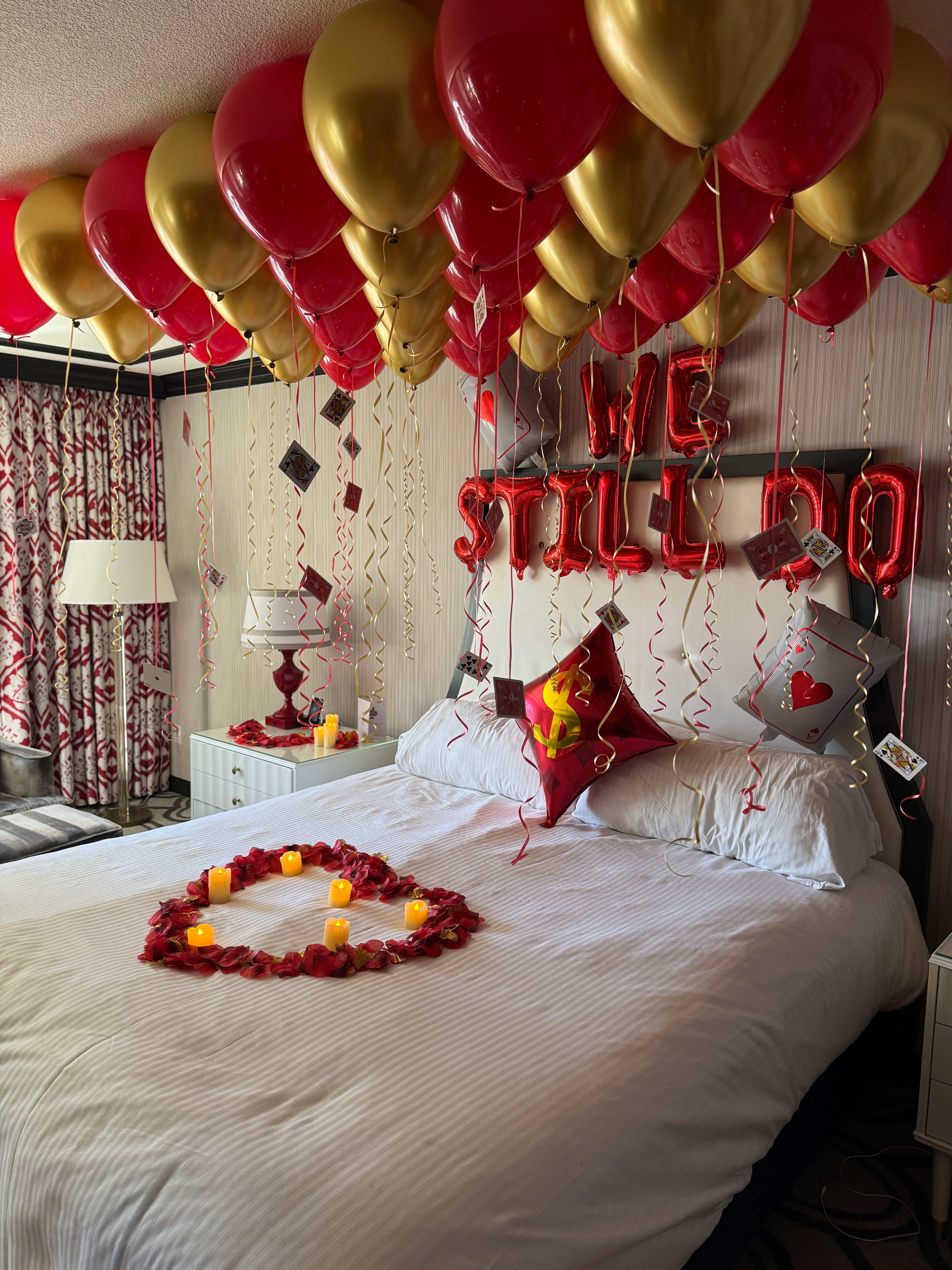 Romantic hotel room decked out with red and gold helium balloons on the ceiling, dangling playing cards and curly ribbons, red foil letter balloons above the headboard, a heart of rose petals on the bed and flickering LED candles.