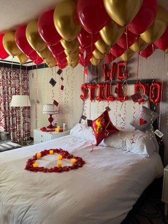 Romantic hotel room decked out with red and gold helium balloons on the ceiling, dangling playing cards and curly ribbons, red foil letter balloons above the headboard, a heart of rose petals on the bed and flickering LED candles.