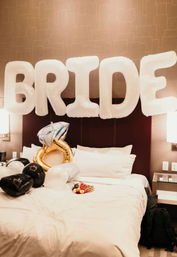 Hotel bridal-suite bed decorated with large white 'BRIDE' balloons, a gold engagement-ring balloon, black and white heart balloons, and a small plate of fruit on the sheets.