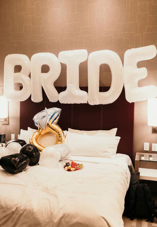 Hotel bridal-suite bed decorated with large white 'BRIDE' balloons, a gold engagement-ring balloon, black and white heart balloons, and a small plate of fruit on the sheets.
