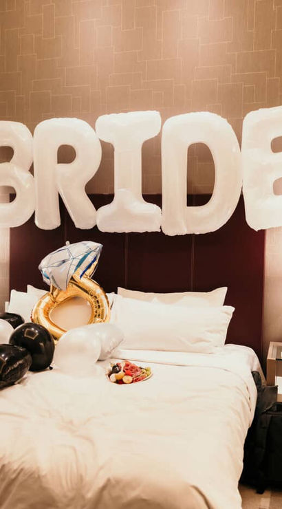 Hotel bridal-suite bed decorated with large white 'BRIDE' balloons, a gold engagement-ring balloon, black and white heart balloons, and a small plate of fruit on the sheets.