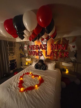 Romantic Las Vegas–themed hotel room surprise with red, black and white balloons, foil Vegas letter balloons, a rose-petal heart and glowing candles on the bed.