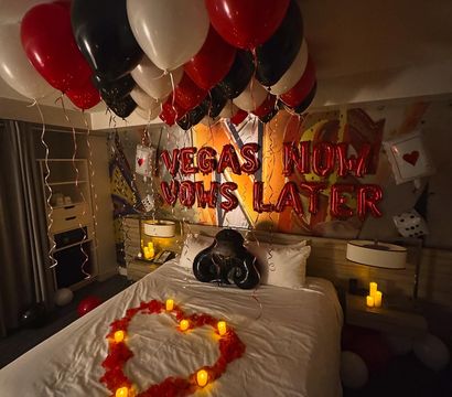 Romantic Las Vegas–themed hotel room surprise with red, black and white balloons, foil Vegas letter balloons, a rose-petal heart and glowing candles on the bed.