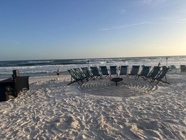 Cozy semicircle of teal lounge chairs around a fire pit on white-sand beach with rolling ocean waves and a clear sunset sky
